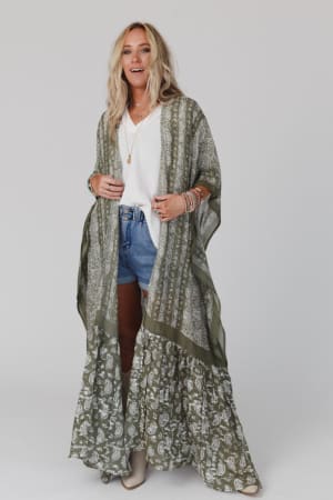 A woman with blonde hair wears the Paisley Tapestry Free Flow Duster Kimono in Olive over a white top and denim shorts, accessorized with layered necklaces and bracelets, standing against a plain white background.