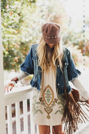 A woman stands outdoors in the Scottie Ruffle Denim Jacket - Blue with ruffled sleeves and flap pockets, a white embroidered dress, a fringed brown bag, and a brown hat. Her long blonde hair flows as she looks down.