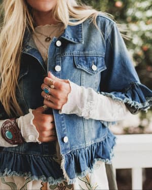 A woman with long blonde hair stands outdoors by greenery, wearing the Scottie Ruffle Denim Jacket - Blue over a white long-sleeve shirt, paired with a flowery skirt and accessorized with rings and a turquoise bracelet.