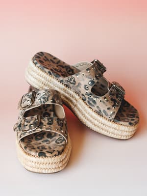 A pair of leopard print platform slide sandals with double buckle straps, silver grommet studding, and a woven raffia-wrapped platform sole, shot against a warm blush pink gradient background.