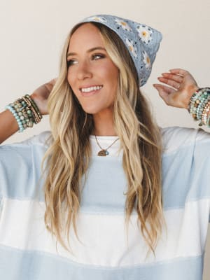 A smiling woman adjusts a powder blue crochet daisy beanie with both hands, styled with a wide-stripe light blue and white top, layered beaded bracelets, and a delicate charm necklace — the whole look gives off easy, feel-good boho energy.