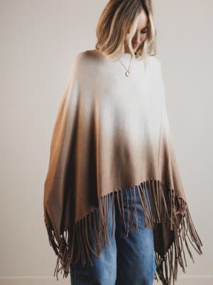Woman wearing a brown and beige gradient poncho with fringe details on a plain background