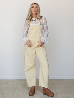 A relaxed, sun-faded vibe — wide-leg cream overalls layered over a sheer lace long-sleeve top, styled with brown clogs and turquoise jewelry for that effortless boho-meets-vintage feel.