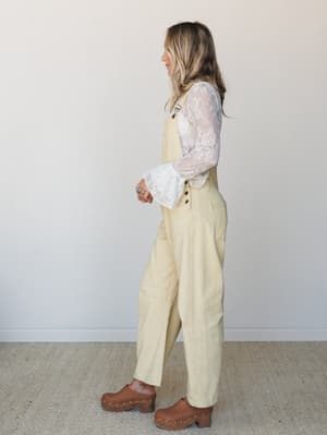 A side-profile shot of a model in buttery yellow overalls layered over a sheer floral lace long-sleeve top, finished with brown wood-studded clogs — the kind of outfit that feels effortless but clearly thought through.