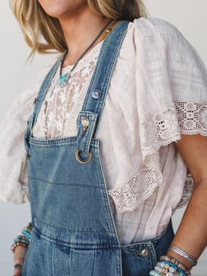A close-up lifestyle shot of denim overalls layered over a blush lace-trim top, styled with turquoise beaded bracelets and a turquoise pendant necklace — the kind of effortless, lived-in combo that looks like it came together naturally.