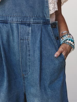 A close-up detail shot of medium-wash denim overalls with a hand tucked into the side pocket, layered with an armful of beaded and silver bracelets — the kind of effortless, lived-in look that feels equally at home on a farm or at a farmers market.
