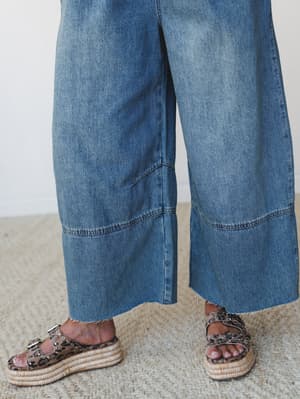 A close-up detail shot of wide-leg denim overalls with raw hem and panel seaming, styled with leopard print platform espadrille sandals on a woven jute rug — the kind of effortless pairing that makes a casual outfit feel thought about.