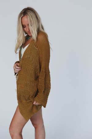 A woman with long blonde hair wears the Brenden Knit Netted Cardigan in brown over a white outfit, standing against a plain light gray background and looking down while holding one side of the cardigan.