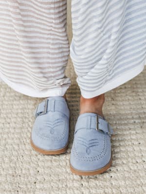 A close-up foot shot of dusty blue suede mules with studded detailing and buckle straps, paired with soft striped wide-leg pants on a chunky woven rug — relaxed and quietly put-together.