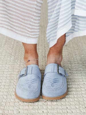A close-up of periwinkle suede mules with silver buckle hardware and brogue-style stitching detail, styled with a striped knit midi skirt and a delicate evil eye anklet — effortless and a little personal.