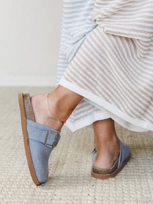A close-up shot of periwinkle blue suede mules with silver buckle detail, styled with a striped knit midi skirt and a delicate anklet — easy, intentional, and very much the kind of shoes you wear everywhere for three seasons straight.
