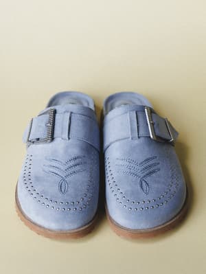 A pair of dusty blue suede mules shot from above against a warm sand background — buckle straps, tonal embroidery, and studded broguing details give them that perfect mix of laid-back and considered.
