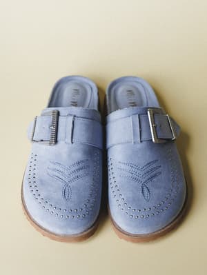 A pair of periwinkle blue suede mules shot overhead on a warm sand background — the kind of shoe that makes you want to wear linen and wander somewhere slow.