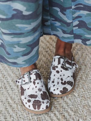 A close-up shot of cow print mules with studded detailing and buckle straps, styled with wide-leg camo pants in blue tones — two bold prints doing exactly what they want on a woven jute rug.