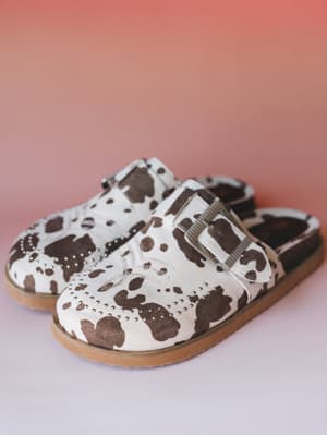 A pair of cow-print slip-on mules with silver stud trim and a ribbed buckle strap, shot against a warm coral-to-lavender gradient — equal parts cozy and personality-forward.