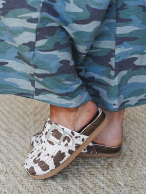 A close-up detail shot of cow-print mule slides paired with a flowing blue camo maxi skirt — the kind of pattern-mixing that feels accidental in the best way.