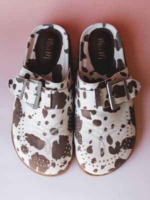 A pair of cow print mule clogs shot overhead on a dusty pink background — cream and brown spots meet silver studded detailing and metal buckle straps for a look that's equal parts western and wearable.