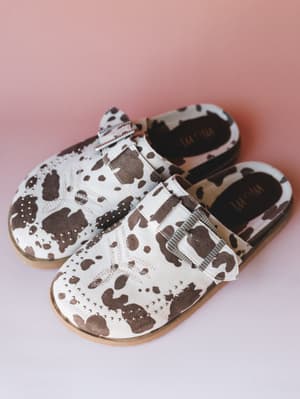A pair of cow print mule clogs with silver stud detailing and a buckle strap, shot against a soft pink backdrop — the kind of shoe that does all the talking so your outfit doesn't have to.