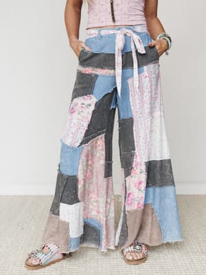 A statement-making pair of ultra-wide-leg patchwork pants stitched together from floral, striped, and washed denim fabrics — worn with a pink crochet crop top and beaded sandals for a free-spirited, artsy-boho vibe.