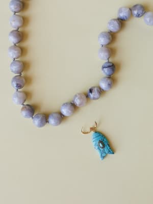 A close-up of a knotted beaded necklace with soft lavender-grey marbled stone beads, finished with a carved turquoise fish charm on a gold hook — the kind of piece that feels like it came home from a coastal antique market.