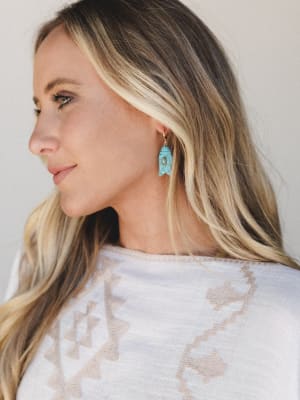 A close profile shot of a model wearing a turquoise stone fish-shaped drop earring with a gold hoop backing, paired with a white top featuring a woven tan geometric/aztec pattern — effortlessly boho and easy to wear.