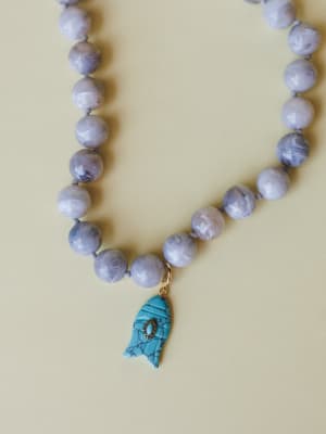 A close-up of a knotted beaded necklace in soft lavender-grey stone beads, centered by a carved turquoise fish charm with gold hardware — the kind of piece that looks like it came home from a coastal market and never left your neck.