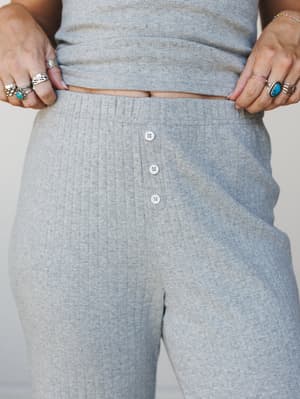 A close-up detail shot of a heather grey ribbed knit matching set — the pants' button-front placket and high waistband take center stage, styled with a stack of silver and turquoise rings that feel very much like a personal collection, not a stylist'