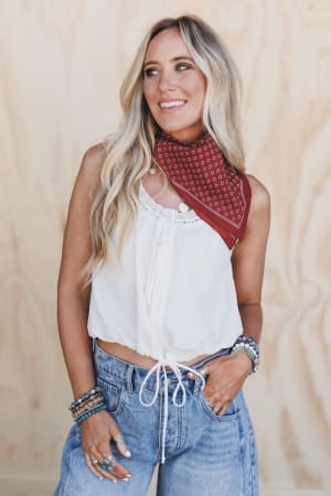A woman with long blonde hair, in a sleeveless white top and blue jeans, smiles with one hand in her pocket, wearing the Rylee Flower Field Bandana Scarf in rust around her neck, standing before a light wood background.