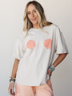 An oversized cream tee with a playful double scallop shell graphic gets styled with blush shorts and a stack of beaded bracelets — casual, coastal, and the kind of thing you'd reach for on a beach-town morning.