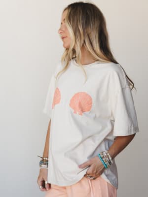 An oversized cream tee with coral scallop shell graphics worn with a stacked bracelet arm and a peek of peachy bottoms — effortless beach-town energy without trying too hard.