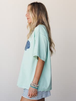 A side-profile shot of a model in an oversized mint tee with a navy seashell graphic, paired with blue and white striped shorts and layered beaded bracelets — effortlessly coastal and easy to reach for.