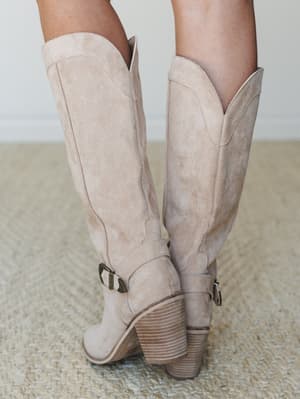 A close-up back view of knee-high suede cowboy-style boots in warm sand beige, showing off the stacked wood heel, ankle buckle hardware, and V-cut topline — the kind of boots that make even a Tuesday feel intentional.