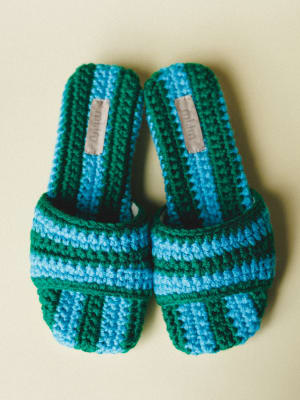 A pair of chunky crochet slide sandals in bold blue and green stripes, shot flat against a warm cream background — the kind of shoes that make you want to pack a beach bag.