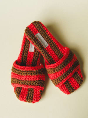 A pair of chunky crochet slide sandals in bold red and brown stripes, shot flat against a soft cream background — the kind of shoes that make you want to pad around the house like it's a lifestyle.