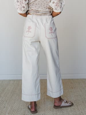 A back-view shot of wide-leg cream pants with charming pink floral embroidery on the patch pockets and contrast stitching at the hem, styled with a lace-sleeved top and strappy sandals — the kind of pants that make a grocery run feel like a story wor