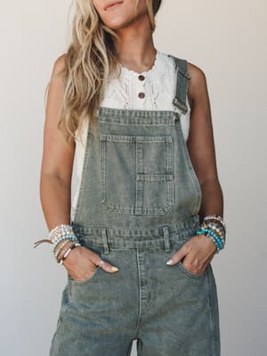 Washed olive denim overalls layered over a cream pointelle knit top, styled with stacks of beaded bracelets — the kind of effortless, lived-in look that feels intentional without trying too hard.