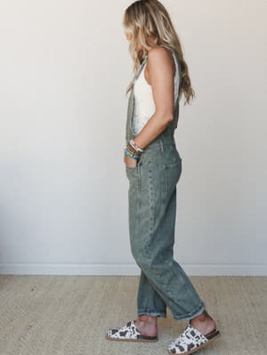 A relaxed side-profile shot of a model in washed olive denim overalls layered over a cream crochet tank, styled with cow-print mules and a stack of colorful beaded bracelets — the kind of effortless outfit that looks like it came together on a good m