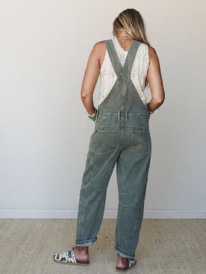 Shot from behind, she's wearing washed olive denim overalls over a cream pointelle tank, styled with cow print slides and stacked bead bracelets — the kind of effortless outfit that looks like it came together in two minutes but somehow feels complet