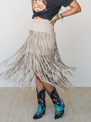 A fringe midi skirt in dove tan suede-like fabric swings mid-movement, styled with an acid wash floral graphic tee knotted at the waist, bold embroidered cowboy boots in turquoise and black, and a stack of colorful beaded bracelets — the whole look h