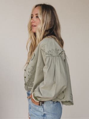 A woman with long blonde hair stands sideways, wearing the Extra Touch Embroidered Denim Jacket in olive with puffed sleeves and blue scalloped-hem jeans. She looks relaxed, accessorizing with beaded bracelets against a plain background.