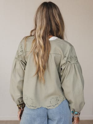 A woman with long, wavy blonde hair is shown from behind, modeling the Extra Touch Embroidered Denim Jacket in olive with balloon sleeves and blue jeans. She stands indoors against a plain beige wall.