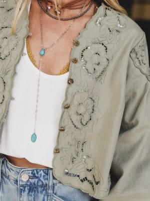 A person wears the Extra Touch Embroidered Denim Jacket in olive, styled with a white tank top, yellow lace bralette, layered necklaces, and high-waisted blue jeans. Only the torso is visible.