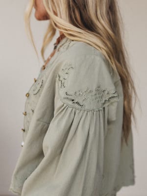 A woman with long, blonde hair models the Extra Touch Embroidered Denim Jacket in olive, featuring embroidered details and a scalloped hem on the sleeves and shoulders, photographed from the side against a neutral backdrop.