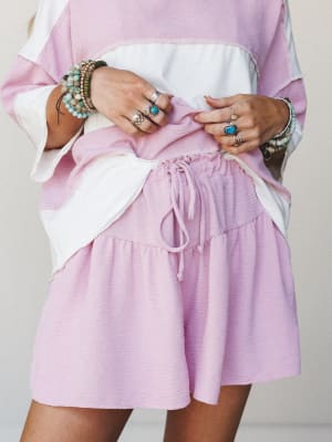 A close-up styled shot of a soft orchid pink and ivory color-block matching set — oversized top with wide sleeves paired with tiered drawstring shorts — hands styled with layered silver rings and beaded boho bracelets pulling the hem, giving it that