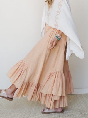 A flowing peach maxi skirt with tiered ruffle hem catches mid-stride, styled with an embroidered white poncho top and strappy sandals — soft, unhurried, and completely wearable.