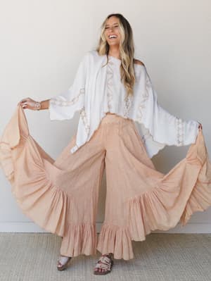 A laughing model fans out the dramatic ruffle hem of ultra-wide-leg pants in soft coral, paired with a flowy white embroidered top — the whole look feels like it belongs somewhere between a beach town market and a backyard dinner with good music.