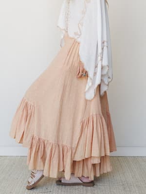 A dreamy side-profile shot of a tiered ruffle maxi skirt in soft peach-coral gauze, paired with a flowing white embroidered wrap top and strappy sandals — the whole look feels like a slow Sunday with nowhere to be.