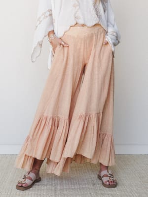 Flowy wide-leg pants in a soft peach-coral with ruffle hem details, styled with a white embroidered top and flat sandals — the kind of outfit that feels as easy as it looks.
