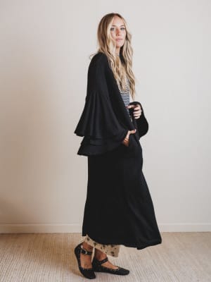 Woman wearing a black duster with a striped top and patterned skirt against a plain background