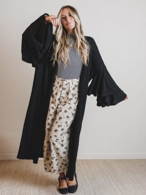 Woman wearing a long black duster over a striped shirt and floral pants indoors.